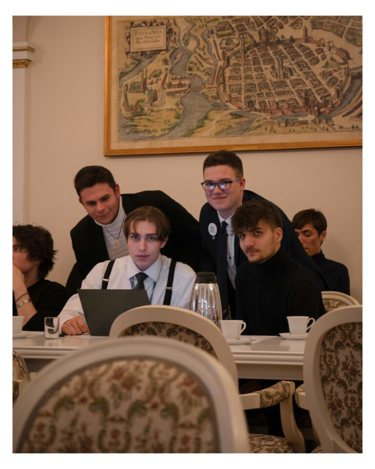 Zdjęcie przedstawia członków Młodzieżowej Rady spoglądających na laptopa podczas sesji Młodzieżowej Rady Miasta Poznania odbywającej się w Sali Białej Urzędu Miasta Poznania przy Placu Kolegiackim 17. W oddali, na ścianie widoczne są historyczne plany Poznania.